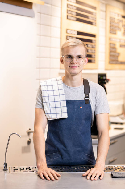 Amical jeune blonde mâle barista avec serviette sur l'épaule debout près du lieu de travail en face de la caméra et vous regardant
 - Photo, image