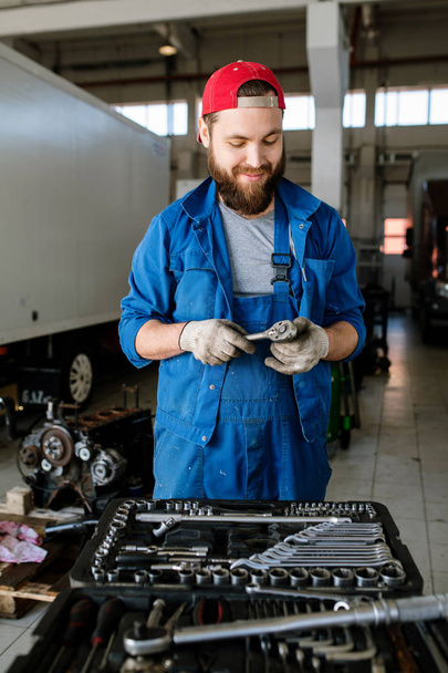 Współczesny młody pracownik serwisu naprawy samochodów stojący przy silniku samochodowym i wybierający narzędzie ręczne - Zdjęcie, obraz