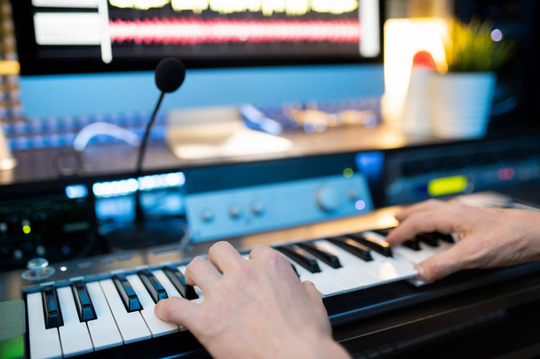 音楽を録音しながら、マイクとコンピュータモニタの前でピアノキーボードのキーを押す若い音楽家の手 - 写真・画像