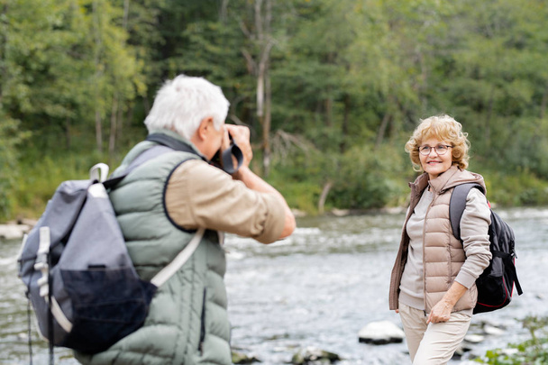 Reifer aktiver Backpacker fotografiert seine glückliche Frau am Ufer des Flusses am Wasser in natürlicher Umgebung - Foto, Bild