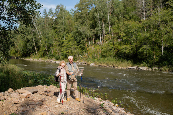 Aktif giyimli yaşlı çift Forest River 'ın yanında duruyor ve yol gösterici harita kullanıyorlar. - Fotoğraf, Görsel