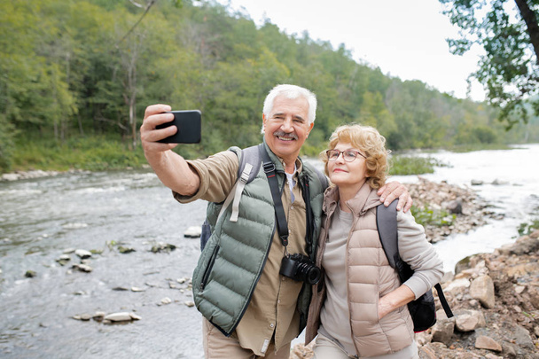 Happy mature active spouses looking at smartphone camera while making selfie by forest river during trip - Фото, зображення