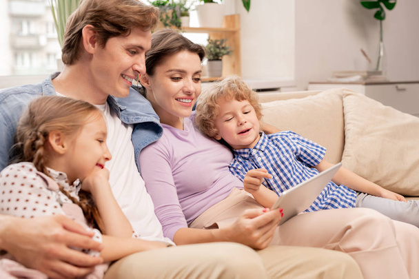 Lindo hijo joven apuntando a la pantalla de la tableta mientras ve dibujos animados con sus padres y hermana en casa por la noche
 - Foto, imagen
