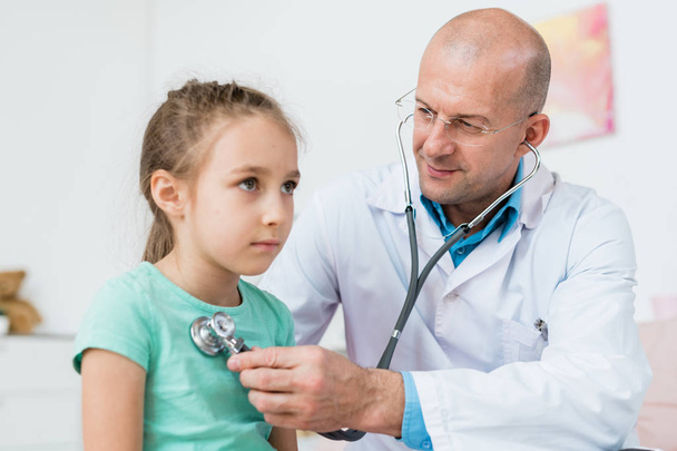 Молодой лысый врач в очках и белом халате, использующий стетоскоп для лечения больной девочки в больнице
 - Фото, изображение