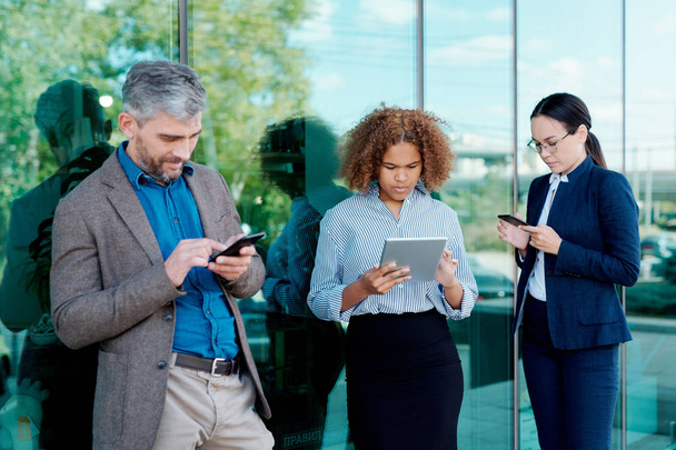 Drie jonge hedendaagse collega 's in formele kleding staan klaar bij kantoorgebouw terwijl ze scrollen in mobiele gadgets - Foto, afbeelding