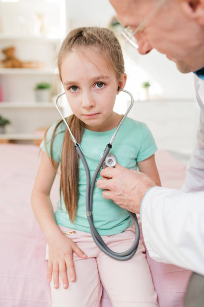 Спантеличена дівчинка слухає серцебиття, в той час як лікар тримає стетоскоп близько до грудей під час огляду
 - Фото, зображення