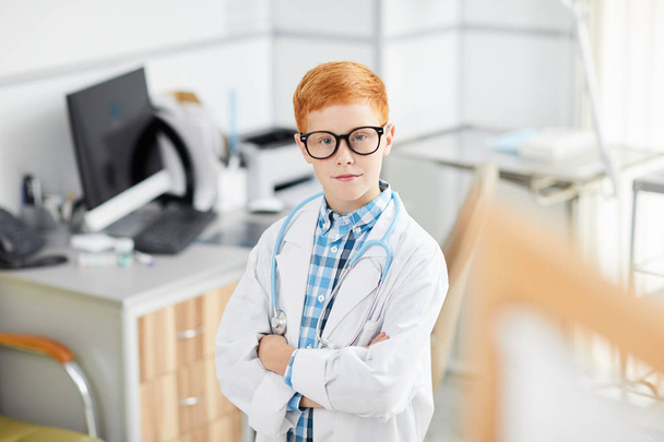 High angle portrait of cute red haired boy posing as doctor wearing white coat while standing with arms crossed and looking at camera confidently, copy space - Valokuva, kuva