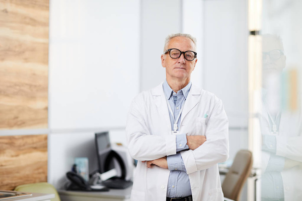 Derékig érő portré idősebb orvos visel szemüveg nézi a kamera, miközben áll a karok keresztbe a hivatalban a modern klinika, másolás tér - Fotó, kép