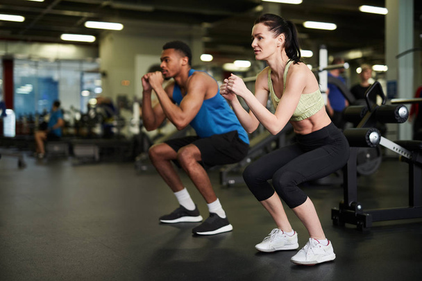 Vue de côté portrait de couple sportif faisant squats pendant l'entraînement de remise en forme dans la salle de gym moderne, espace de copie
 - Photo, image