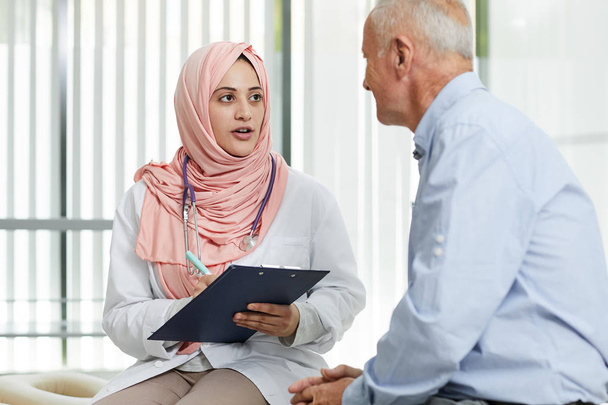 Portret van een jonge Arabische vrouw die werkt als arts in een medische kliniek en praat met oudere patiënten die het formulier invullen op het klembord, kopieerruimte - Foto, afbeelding