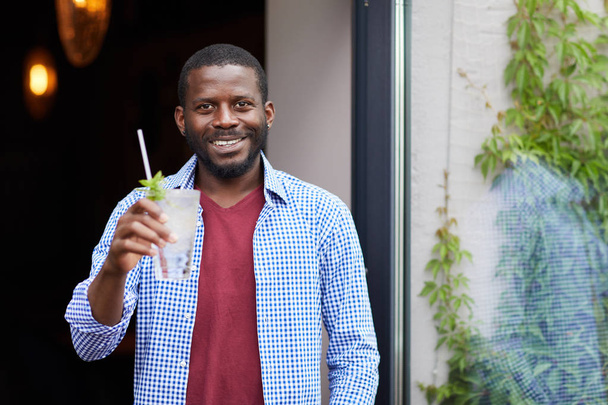 Retrato de la cintura hacia arriba del hombre afroamericano sonriente sosteniendo la bebida fría al aire libre mientras posa en la cafetería, espacio para copiar
 - Foto, Imagen