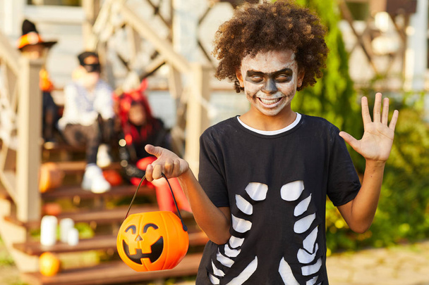 hüfthoch Porträt eines afrikanisch-amerikanischen Jungen im Halloween-Kostüm, der in die Kamera schaut, während er draußen mit einem Kürbiskorb in der Hand posiert - Foto, Bild
