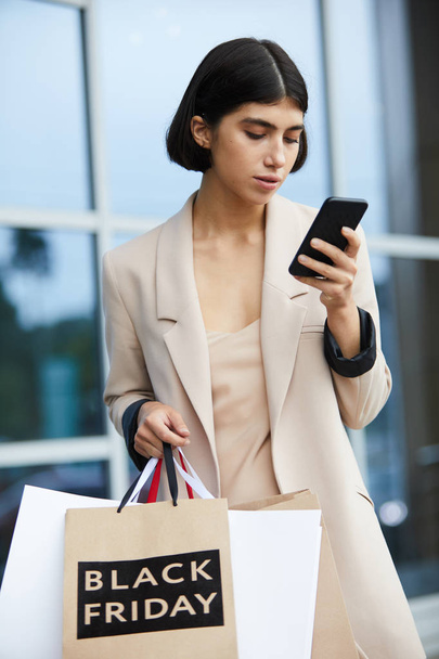 Retrato de la cintura hacia arriba de una mujer joven y elegante sosteniendo bolsas de compras con Black Friday y usando un teléfono inteligente mientras sale del centro comercial en temporada de venta
 - Foto, Imagen