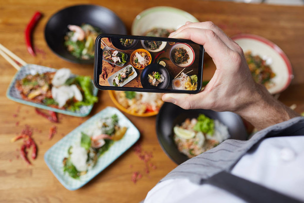 Closeup of unrecognizable man taking photo of delicious Asian food set on wooden table in cafe or restaurant, focus on smartphone screen, copy space - Фото, изображение