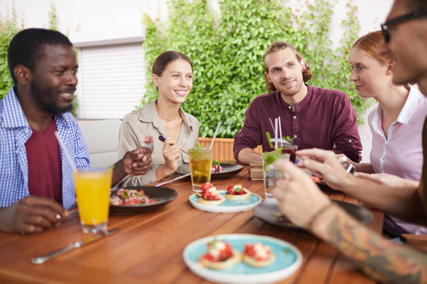 Портрет многонациональной группы друзей, наслаждающихся обедом вместе, сидящих за столом в клетке и счастливо смеющихся, копирующих пространство
 - Фото, изображение