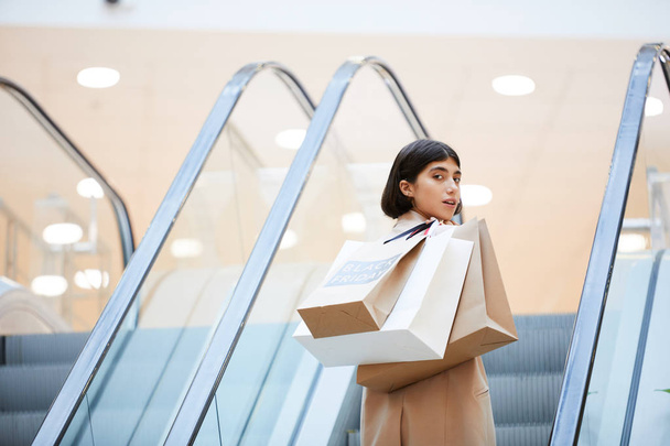 Portrait of beautiful young woman holding shopping bags with Black Friday looking over shoulder at camera while going up escalator in mall, copy space - Photo, Image
