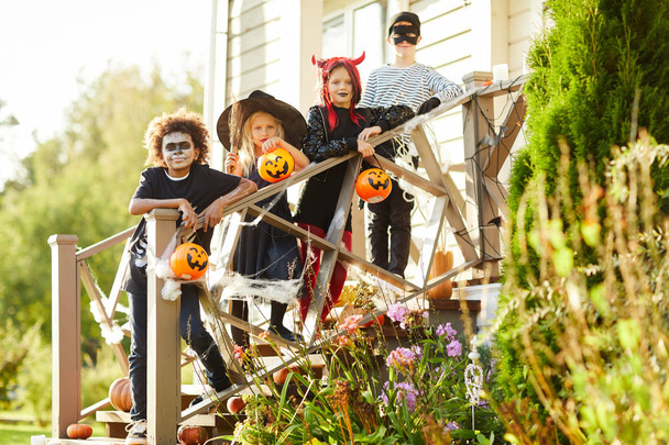 Мульти-этническая группа детей в костюмах на Хэллоуин, стоящих на лестнице украшенного дома, наслаждаясь вместе сладостями или угощениями, копируют пространство
 - Фото, изображение