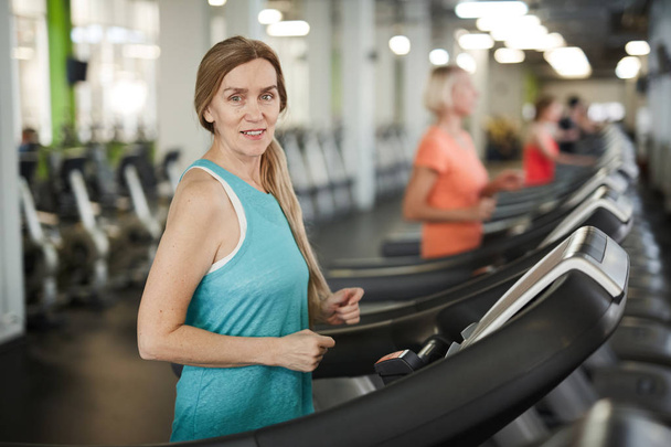 Portrait taille haute de femme mature regardant la caméra tout en courant sur tapis roulant pendant l'entraînement cardio dans la salle de gym moderne, espace de copie
 - Photo, image