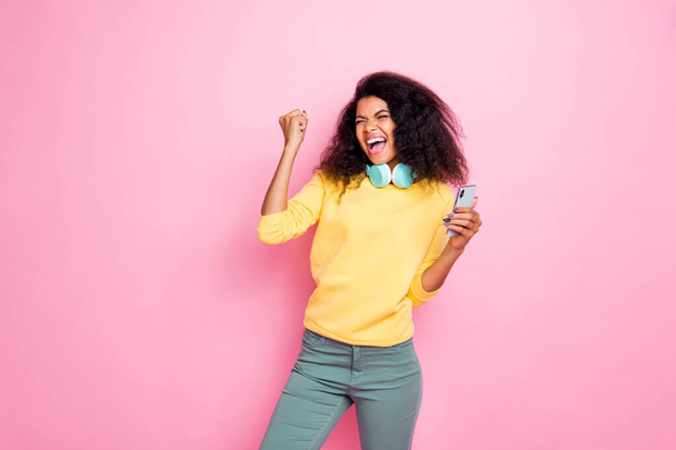 Portrait of crazy funky hipster african girl use cellphone get feedback from followers raise fists scream yes celebrate achievement wear yellow sweater pants isolated over pink color background - Foto, Imagen