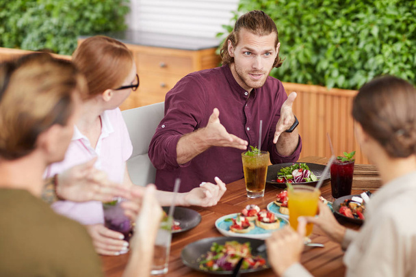Retrato de belo jovem gesticulando ativamente enquanto conversa com amigos sentados à mesa no café durante a reunião de férias, espaço de cópia
 - Foto, Imagem