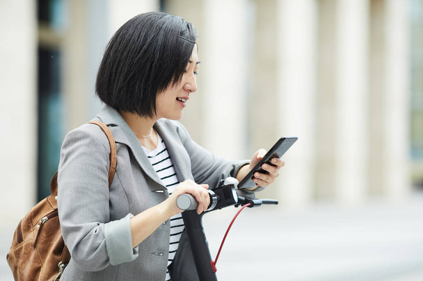 Waist up side view of modern Asian woman checking smartphone while riding electric scooter in urban setting, copy space - Photo, Image