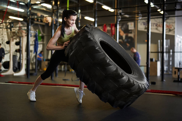 Ganzkörperporträt einer fitten jungen Frau, die während des Krafttrainings im modernen Fitnessstudio einen Reifen umkippt, Kopierraum - Foto, Bild
