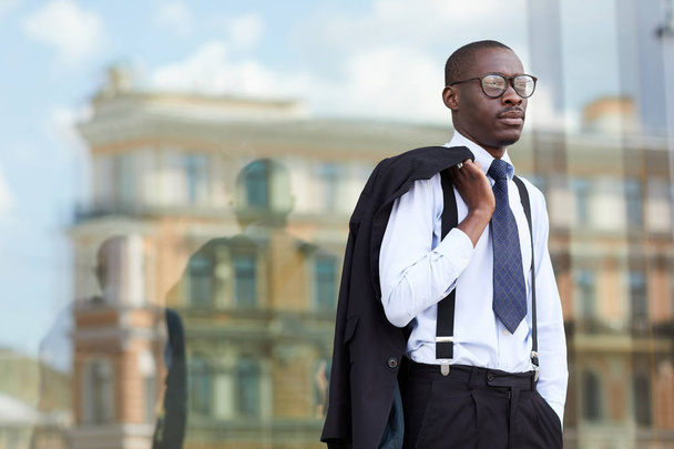 Талия портрет элегантного афро-американского бизнесмена в очках, стоящих на улице у стеклянного окна и держащих куртку, копировальное пространство
 - Фото, изображение