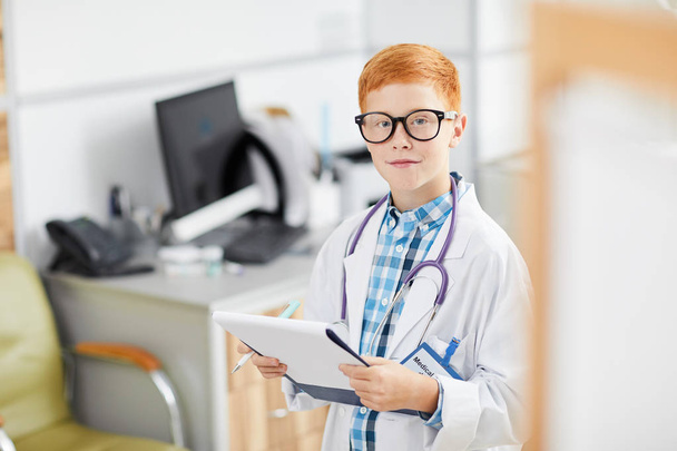 Portrait of cute red haired boy posing as doctor wearing white coat and smiling happily at camera holding clipboard, copy space - Foto, afbeelding