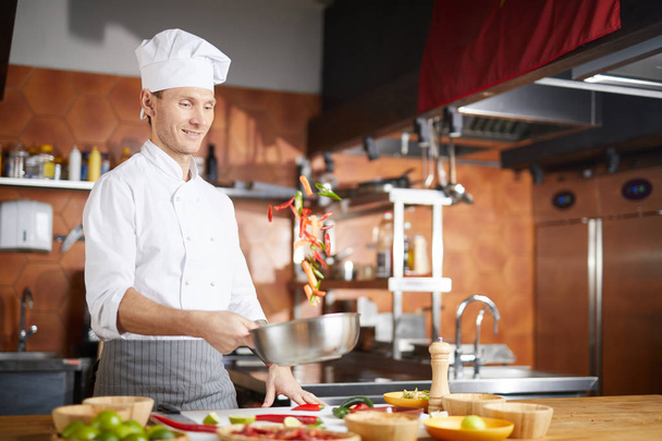 Cintura hacia arriba retrato de guapo chef lanzando verduras mientras cocina plato asiático en la cocina del restaurante, espacio de copia
 - Foto, imagen