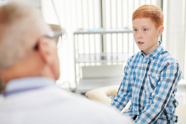 Πορτρέτο του χαριτωμένο έφηβος αγόρι μιλάμε για ανώτερος γιατρός, ενώ κάθεται στον πάγκο κατά τη διάρκεια της διαβούλευσης στη σύγχρονη παιδική κλινική υγειονομικής περίθαλψης, αντίγραφο χώρο - Φωτογραφία, εικόνα