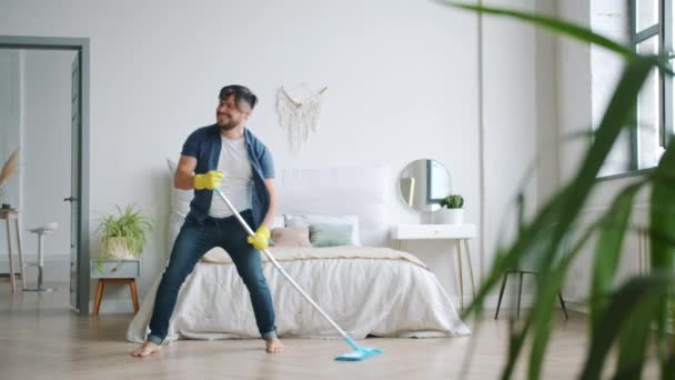 Cheerful young man dancing singing washing floor in apartment with plastic mop - Materiał filmowy, wideo