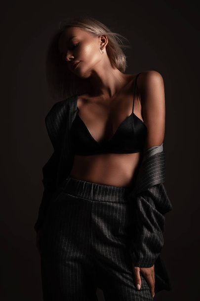 Σέξι ξανθιά κοπέλα με glamour χτένισμα ποζάρουν σε μαύρο φόντο σε εσώρουχα και κλασικό παντελόνι. Πορτρέτο του κομψό σαγηνευτικό καυτό σέξι εσώρουχα κορίτσι στο δωμάτιο - Φωτογραφία, εικόνα