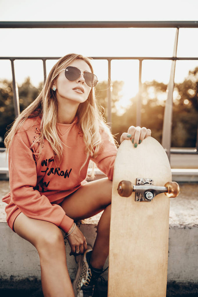 Retrato de beleza de menina ativa com skate na estrada perto de estacionamento no pôr do sol de verão à noite. Modelo de mulher urbana elegante posando em vermelho com capuz, tênis
 - Foto, Imagem