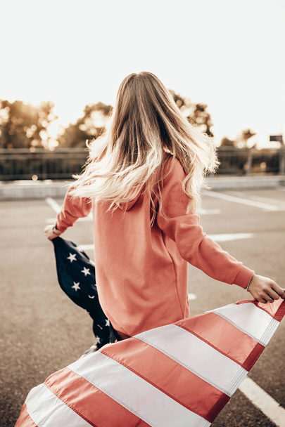 Menina elegante urbana posando com bandeira americana com capuz vermelho no pôr do sol de verão no estacionamento perto da estrada. Retrato ao ar livre modelo de mulher na moda
 - Foto, Imagem