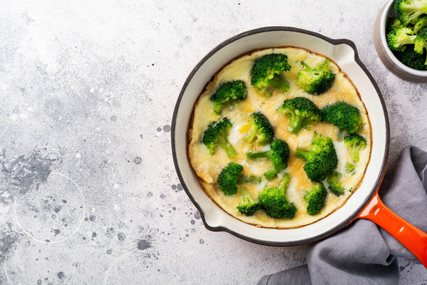 Omelette with broccoli and green young onions on red cast iron pan on old gray concrete background. Top view. - 写真・画像