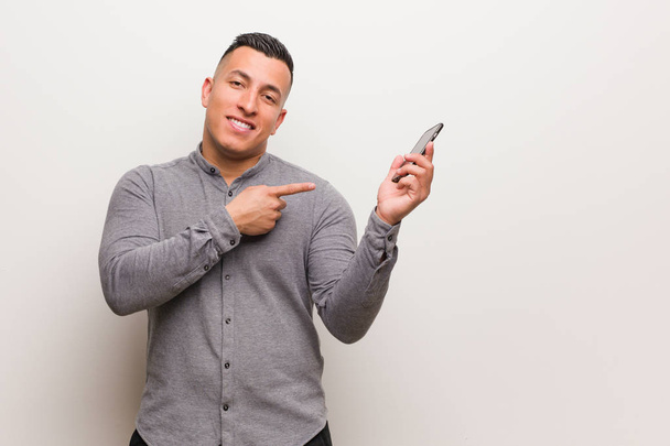 Joven latino sosteniendo un teléfono apuntando hacia un lado con el dedo
 - Foto, Imagen