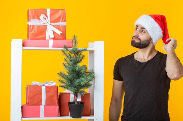 Young positive man with a beard and Santa Claus hat is standing near booth with gifts and Christmas tree on yellow background. Concept of congratulation gift and discounts for Christmas and New Year. - Photo, Image