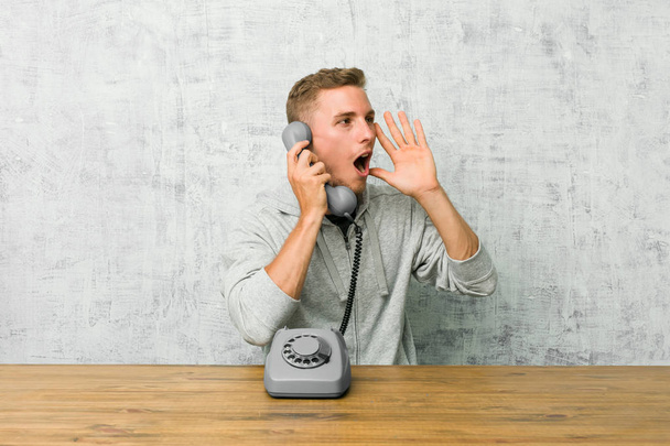Молодой человек, разговаривающий по старинному телефону громко кричит, держит глаза открытыми и руки напряженными
. - Фото, изображение