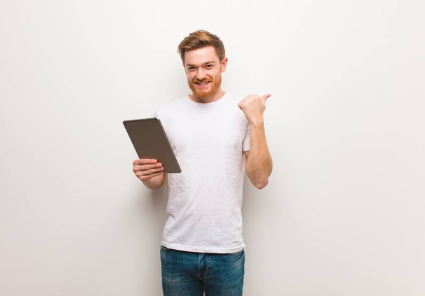 Young redhead man pointing to the side with finger. Holding a tablet. - Photo, Image