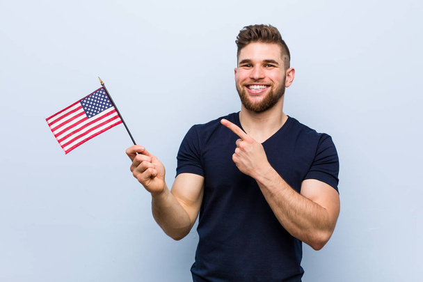 Jovem caucasiano segurando uma bandeira dos Estados Unidos sorrindo alegremente apontando com o dedo indicador de distância
. - Foto, Imagem