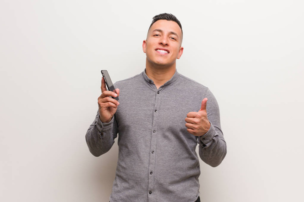 Joven latino sosteniendo un teléfono sonriendo y levantando el pulgar
 - Foto, Imagen