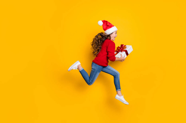 Full length profile photo of amazed small lady jumping high taking giftbox from santa in x-mas midnight wear red knitted sweater and jeans isolated yellow background - 写真・画像