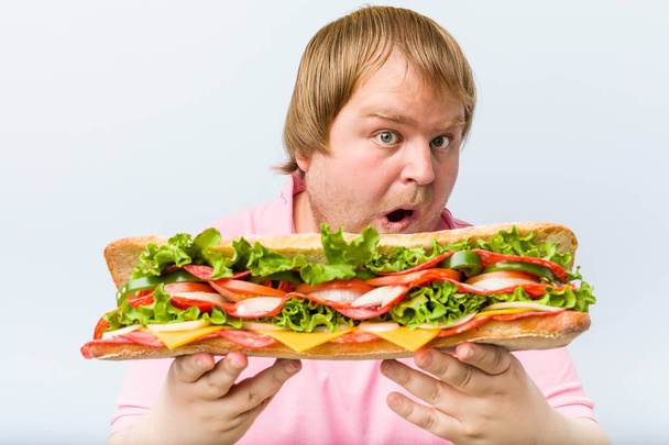 Καυκάσιος τρελός ξανθός χοντρός άνθρωπος κρατώντας ένα τεράστιο σάντουιτς - Φωτογραφία, εικόνα