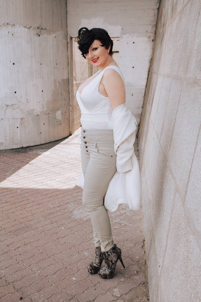 Jonge mooie stijlvolle vrouw in witte wollen jas loopt door de straat. Plus size kort haar volwassen meisje poseren alleen. - Foto, afbeelding