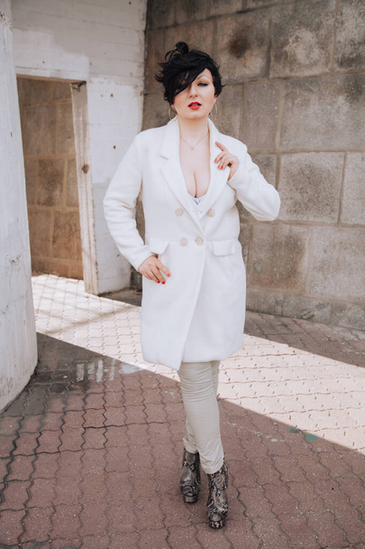 Νέα όμορφη κομψή γυναίκα σε λευκό παλτό μαλλί με τα πόδια κάτω από το δρόμο. Συν μέγεθος κοντά μαλλιά ενηλίκων κορίτσι ποζάρουν μόνοι. - Φωτογραφία, εικόνα