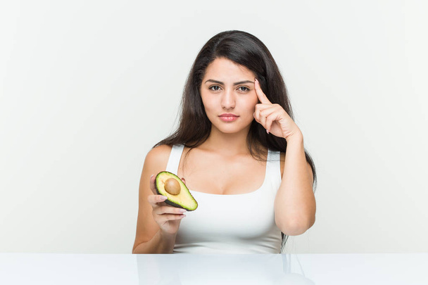 Молодая латиноамериканка держит авокадо, показывая пальцем на висок, думая, сосредоточившись на задаче
. - Фото, изображение