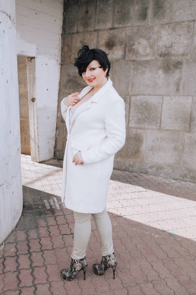 Young beautiful stylish woman in white wool coat walking down the street. Plus size short hair adult girl posing alone. - Photo, Image