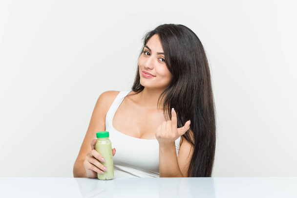 Young hispanic woman holding a vegetable drink pointing with finger at you as if inviting come closer. - Foto, imagen
