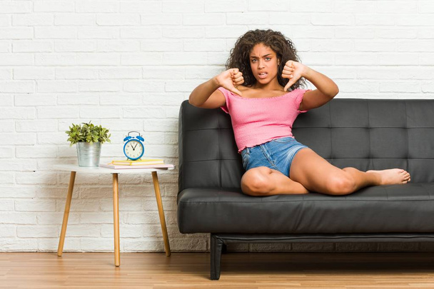 Młoda Afroamerykanka siedząca na kanapie z kciukiem w dół i wyrażająca niechęć. - Zdjęcie, obraz