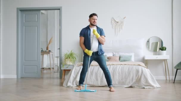 Funny young man singing in mop during clean-up in bedroom having fun alone - Video